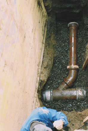 Ein Mann in einer blauen Jacke arbeitet im Dreck an einem Rohr.