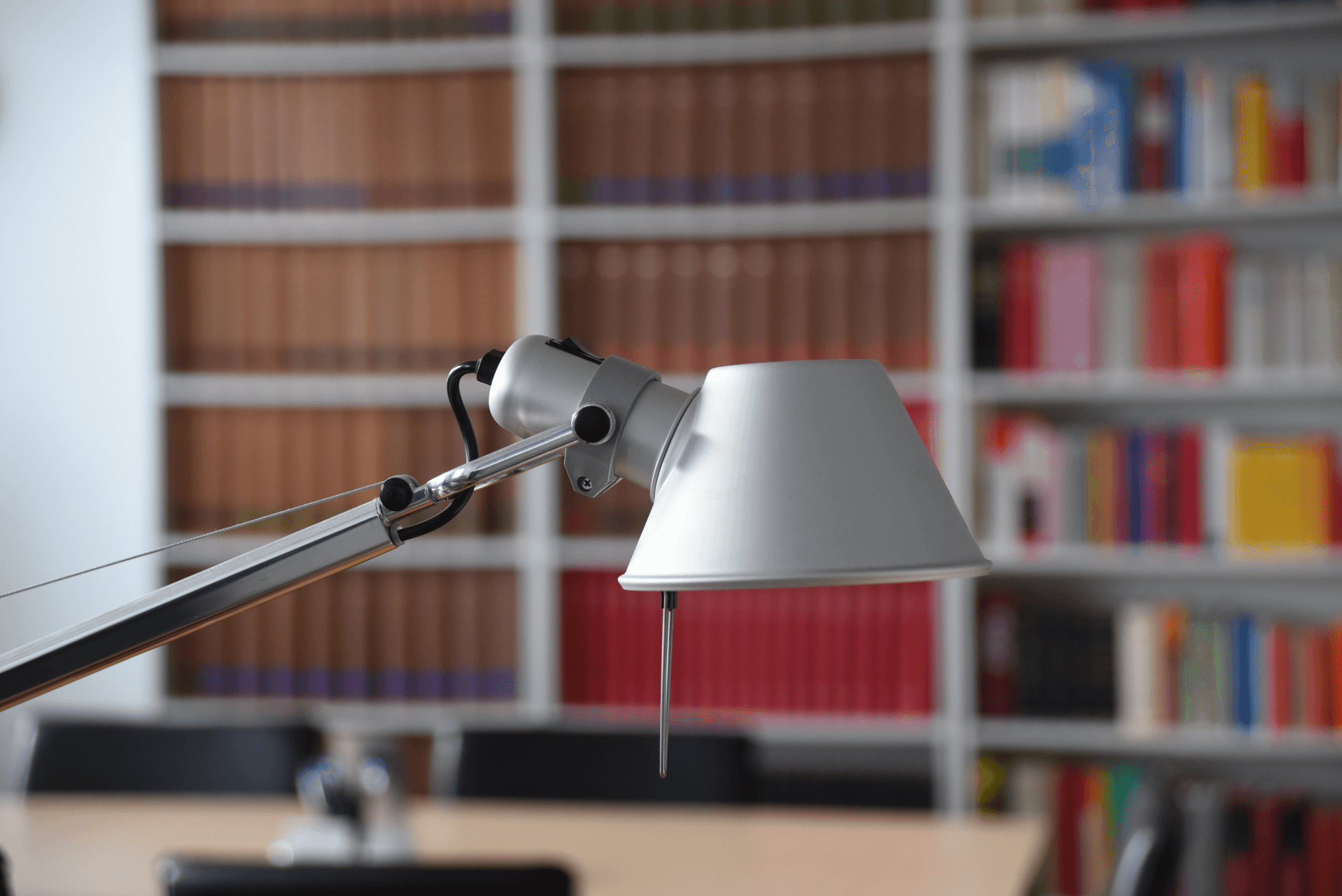 Im Vordergrund eine silberne Schreibtischlampe, im Hintergrund verschwommene Bücherregale aus einer Bibliothek.
