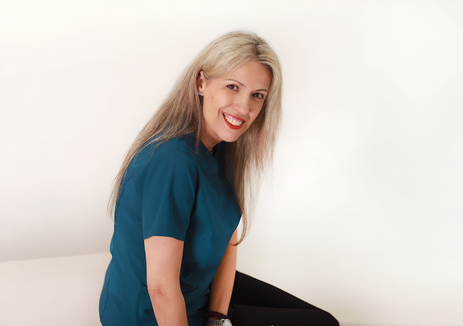 Mujer sonriente con cabello rubio largo, vestida con una camisa color turquesa, sentada contra una pared blanca.