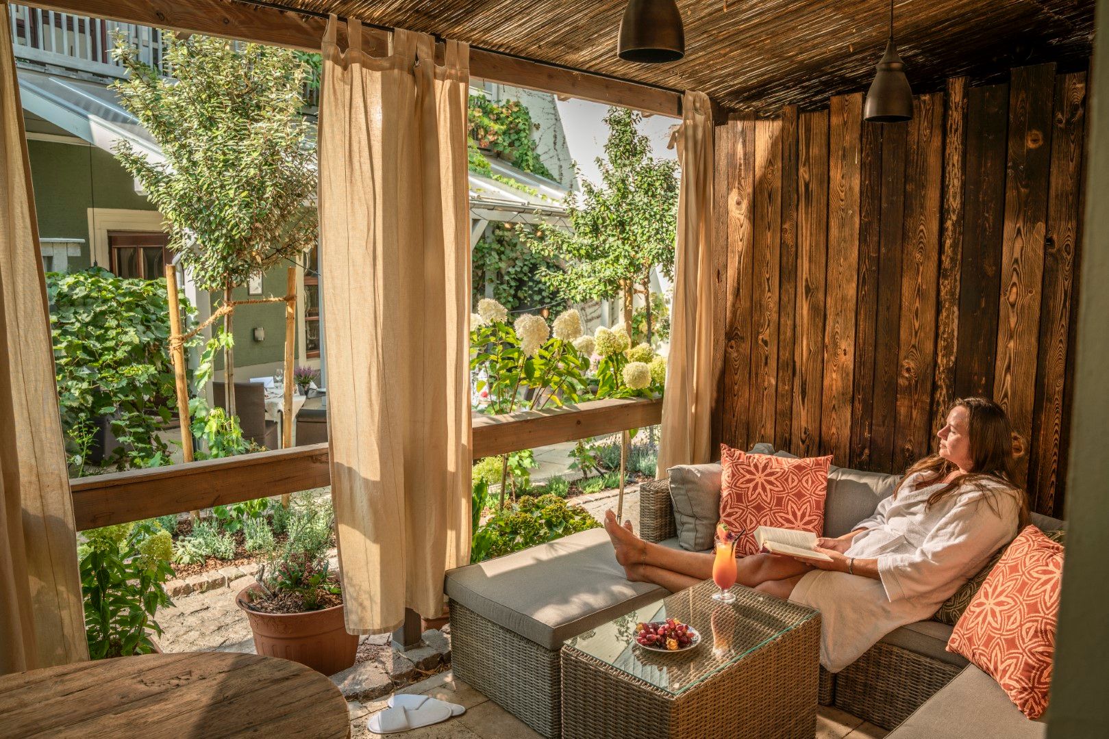 Frau im Morgenmantel entspannt auf einem Sofa im Freien, liest, genießt ein Getränk und den Blick in den Garten.