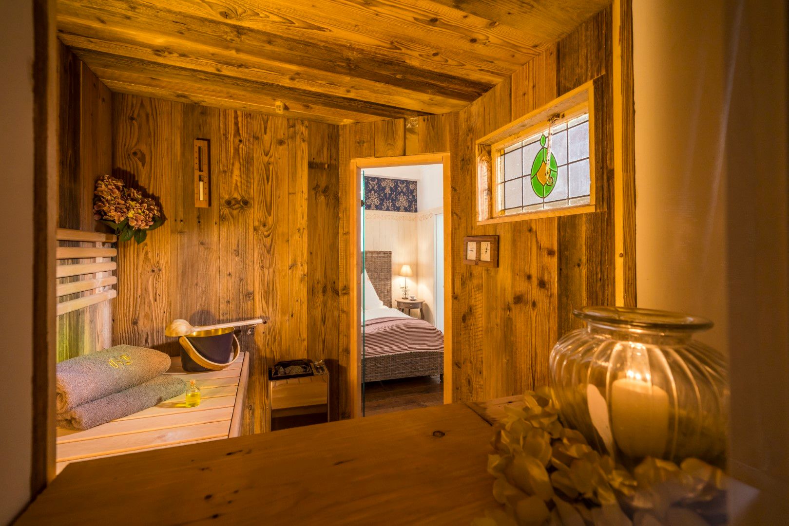 Innenbereich einer hölzernen Sauna mit durch die Tür sichtbarem Bett, beleuchtet von warmem Licht, verziert mit Buntglas.