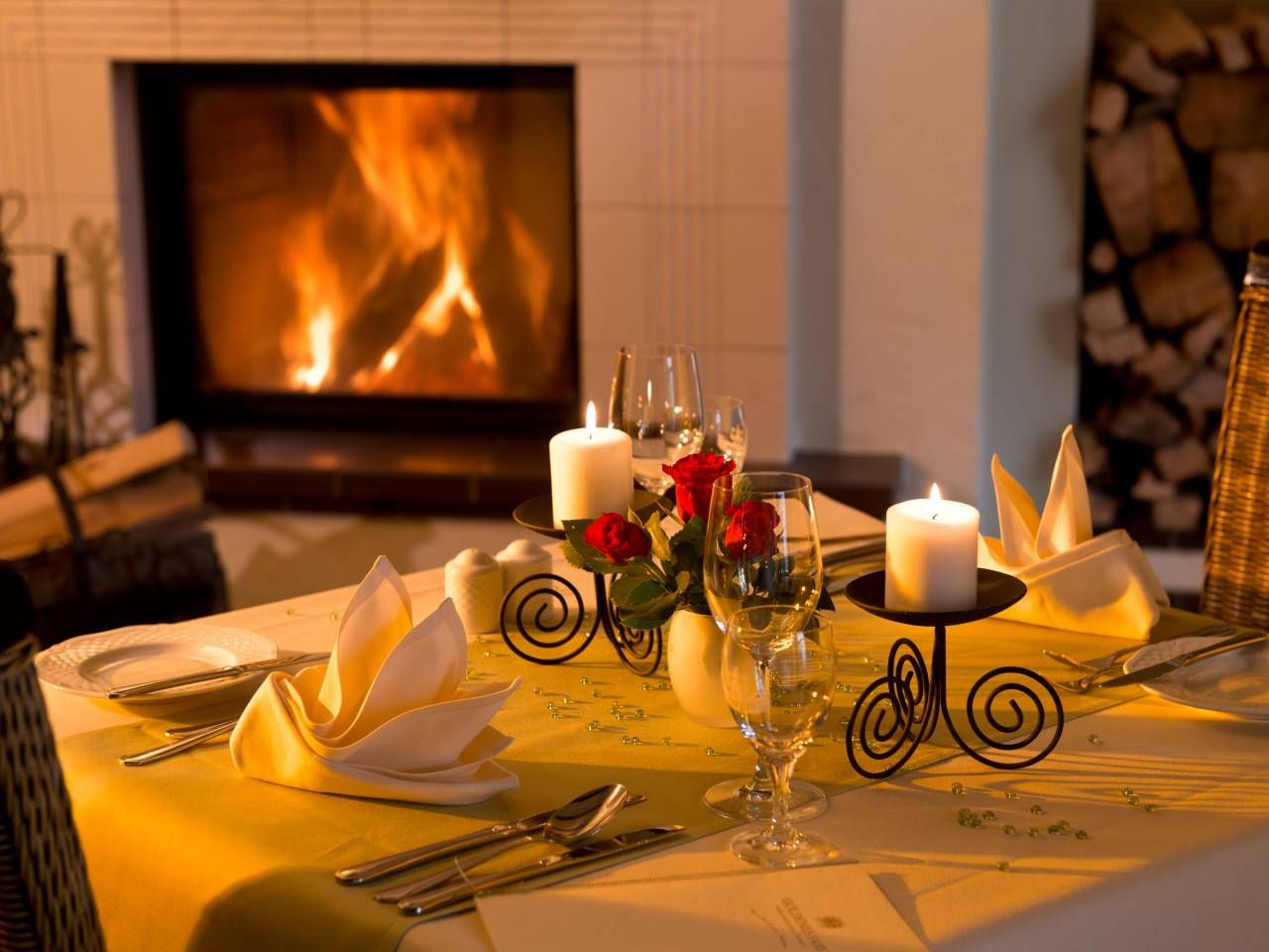 Ein romantisch gedeckter Esstisch vor einem Kamin mit brennenden Kerzen und Rosen.