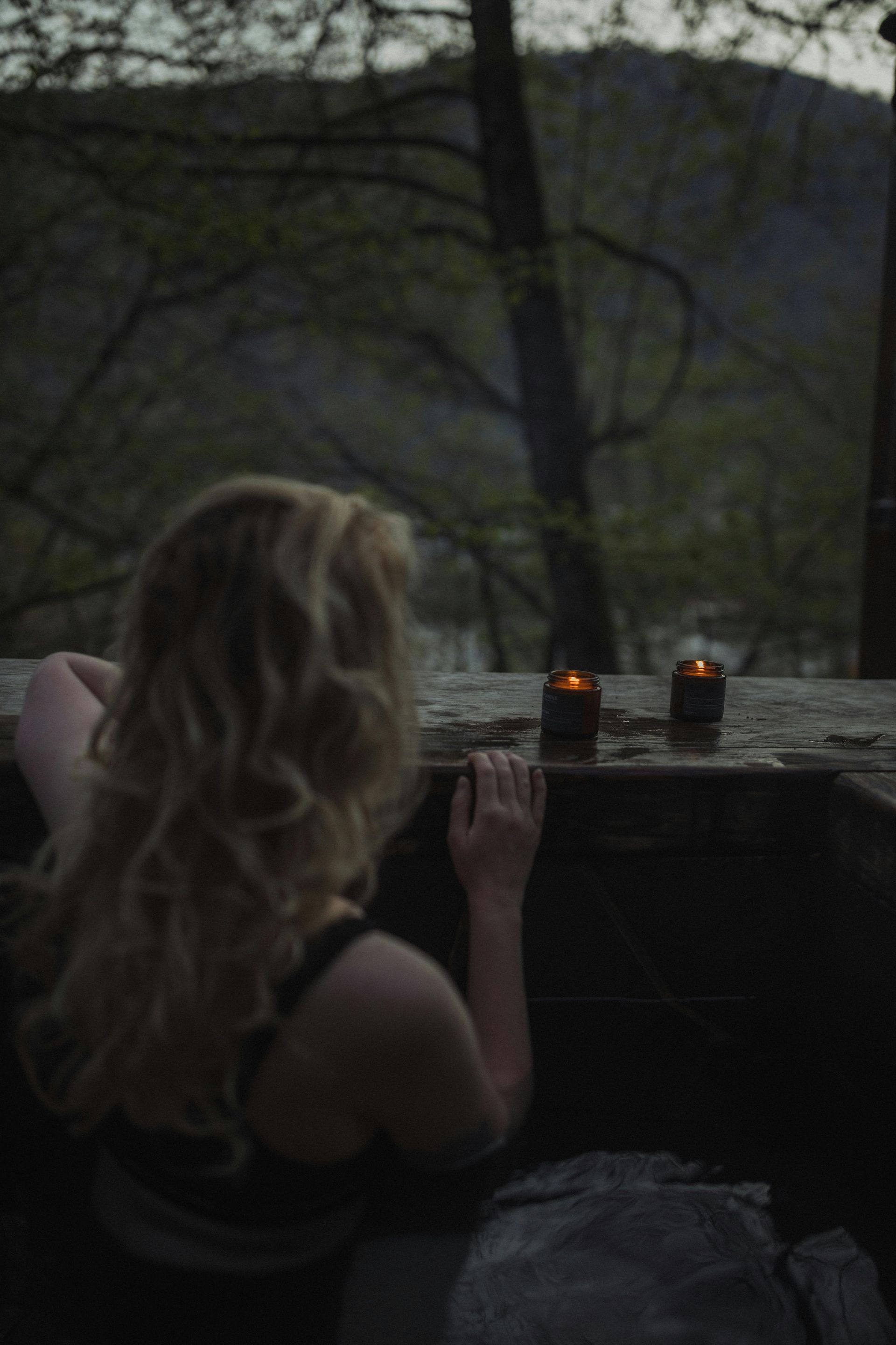 Blonde Person lehnt mit Kerzen an einem Sims; im Hintergrund ein vom Abendlicht erhellter Wald.