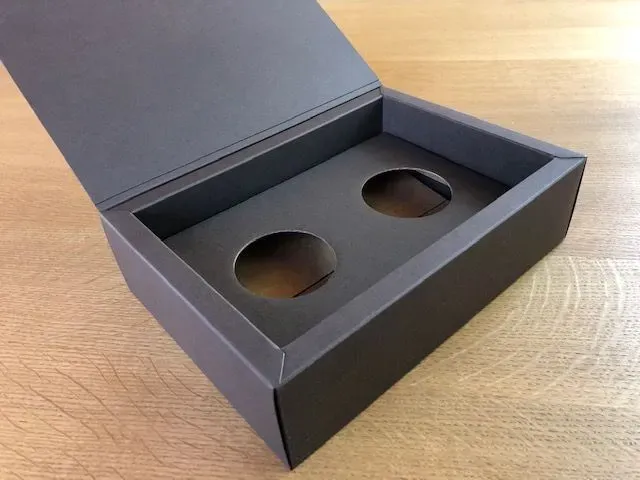 Graue Schachtel mit zwei kreisrunden Ausschnitten, Deckel geöffnet, auf einer Holzoberfläche.