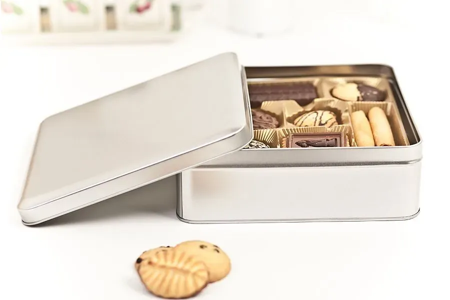 Silberne Dose mit verschiedenen Pralinen, Deckel geöffnet, davor zwei Kekse.