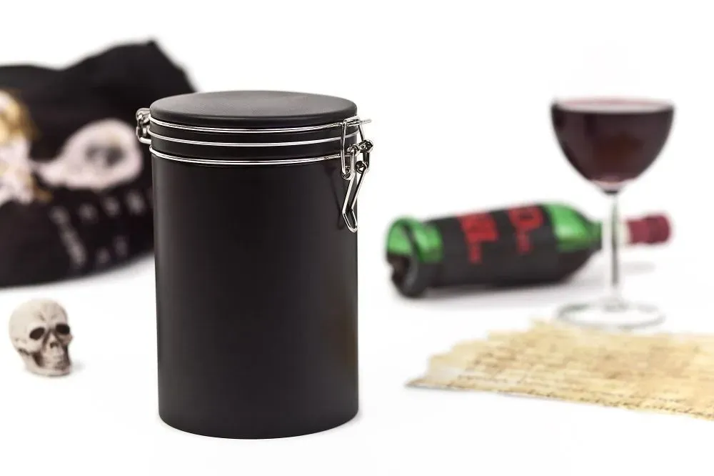 Schwarzer Kanister mit Metallverschluss, Totenkopf, Weinglas und Flasche auf weißem Untergrund.