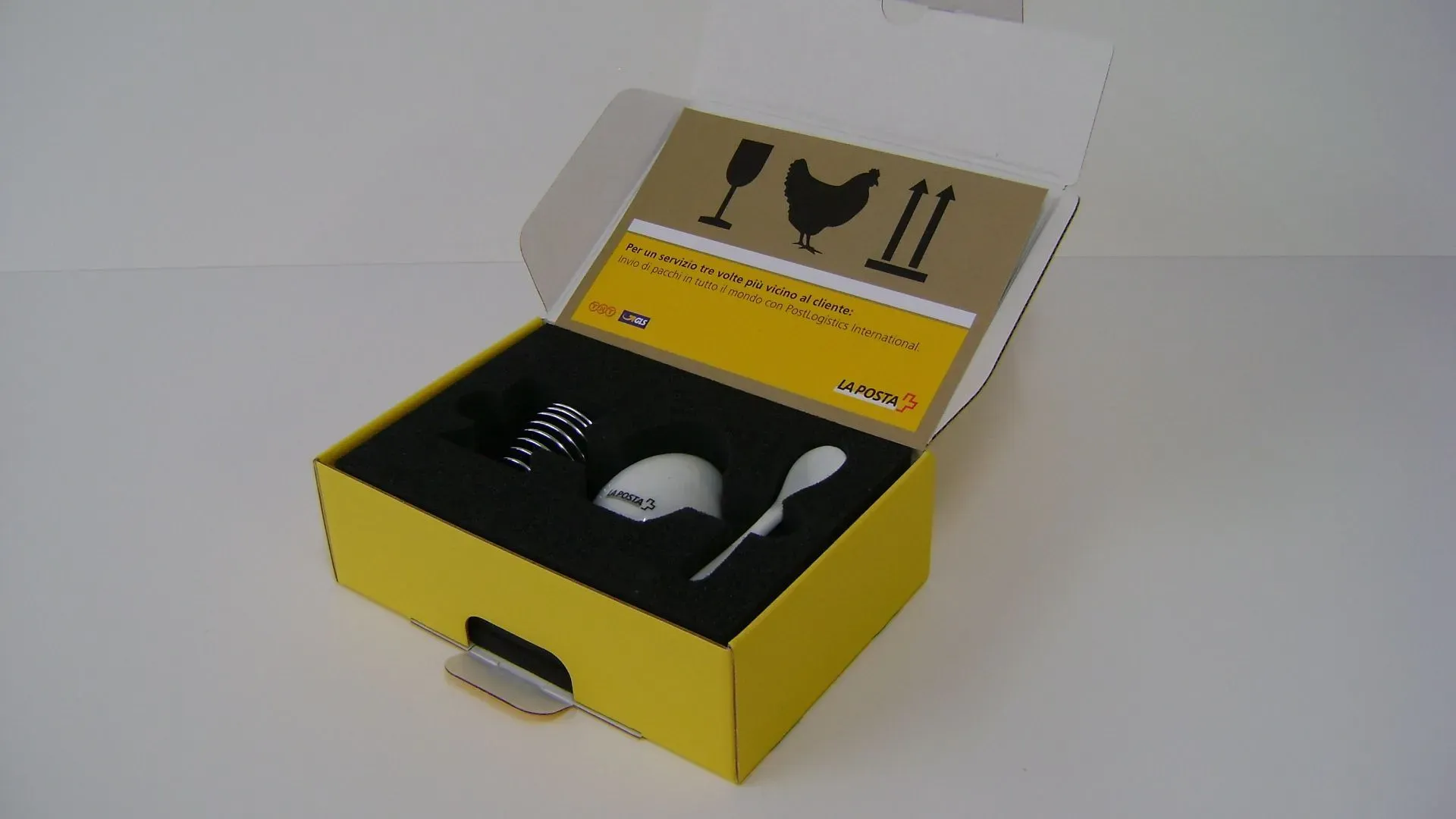 Gelbe Schachtel mit Schaumstoffeinlagen, in der sich ein Eierbecher, ein Löffel und ein Ei befinden; Verpackungshinweise sind sichtbar.