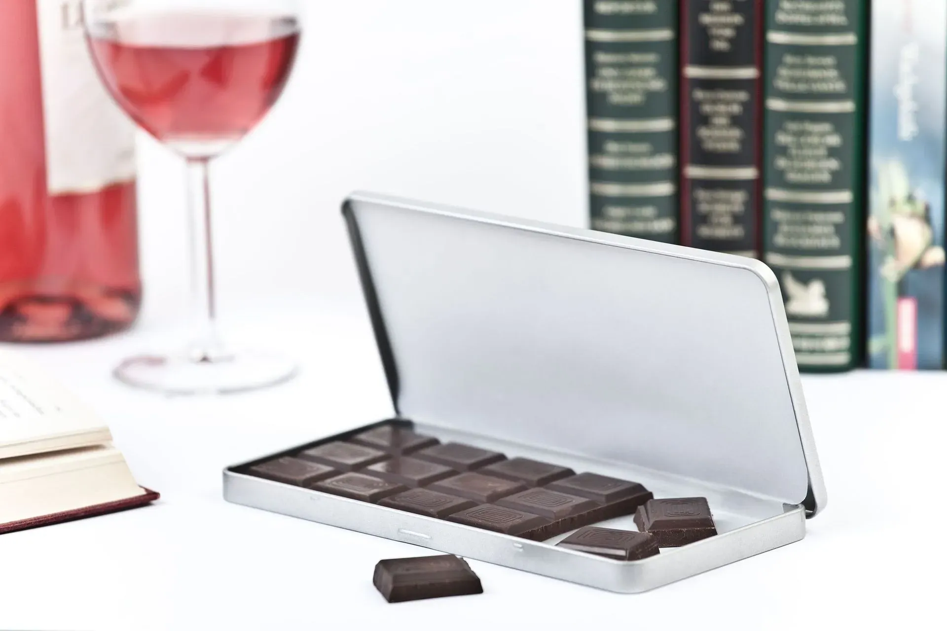 Schokolade in einer Dose, neben Roséwein, Büchern und einem Glas auf einer weißen Oberfläche.