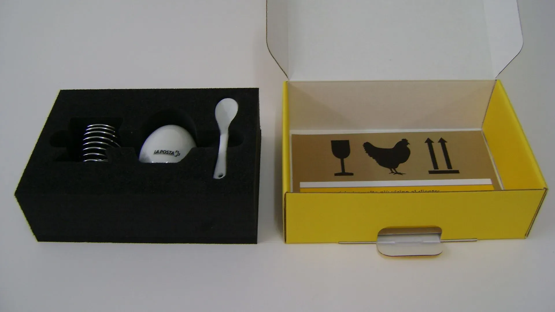 Eierbecher-Set: schwarzer Schaumstoff mit Feder-Eierhalter, weißem Ei und Löffel. Gelbe Schachtel mit Hinweis auf Zerbrechlichkeit.