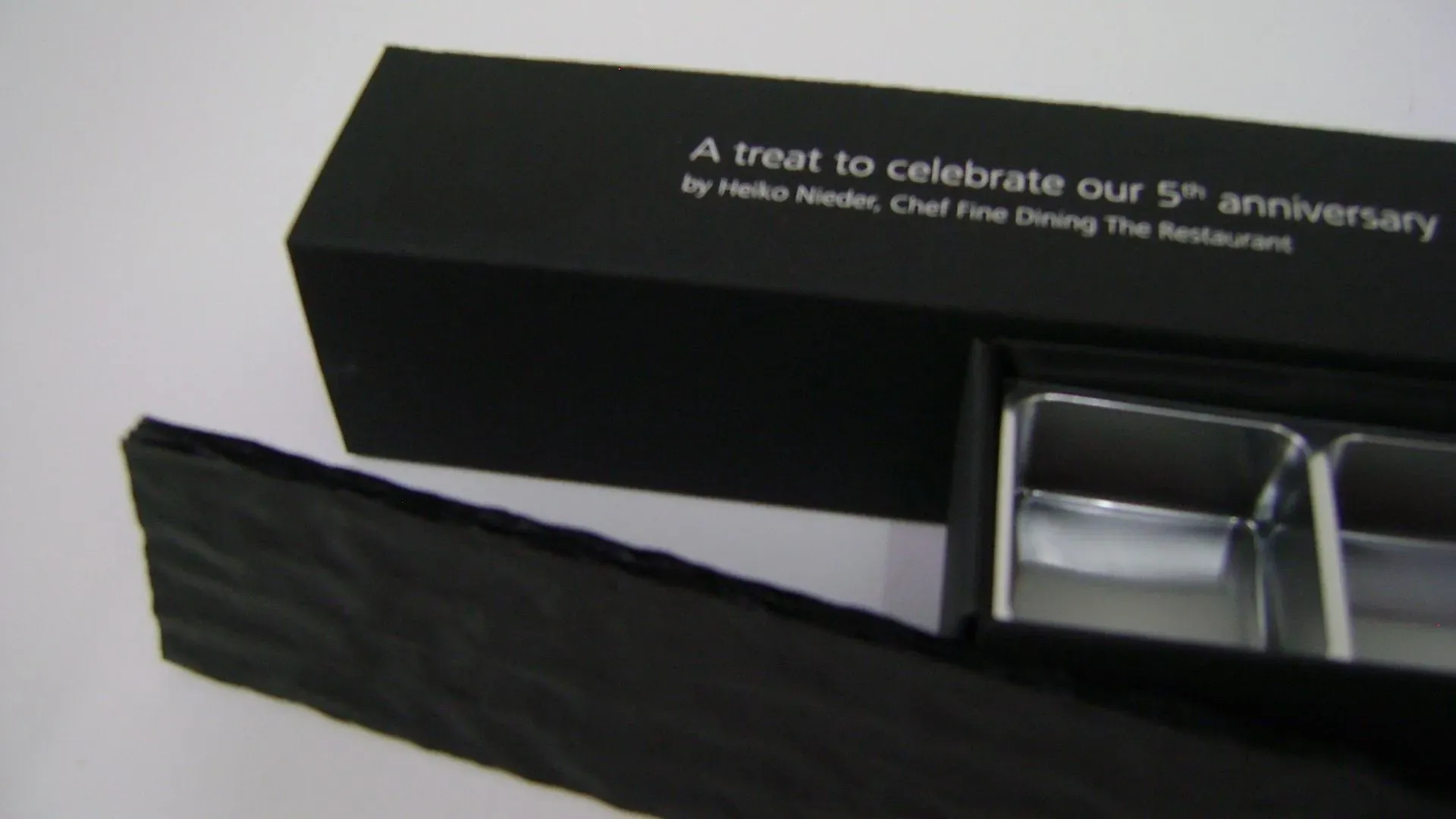 Schwarze Geschenkbox. Beim Öffnen kommen zwei silberne Behälter zum Vorschein. Der Text auf der Box lautet: „Ein Leckerbissen zur Feier unseres 5-jährigen Jubiläums.“