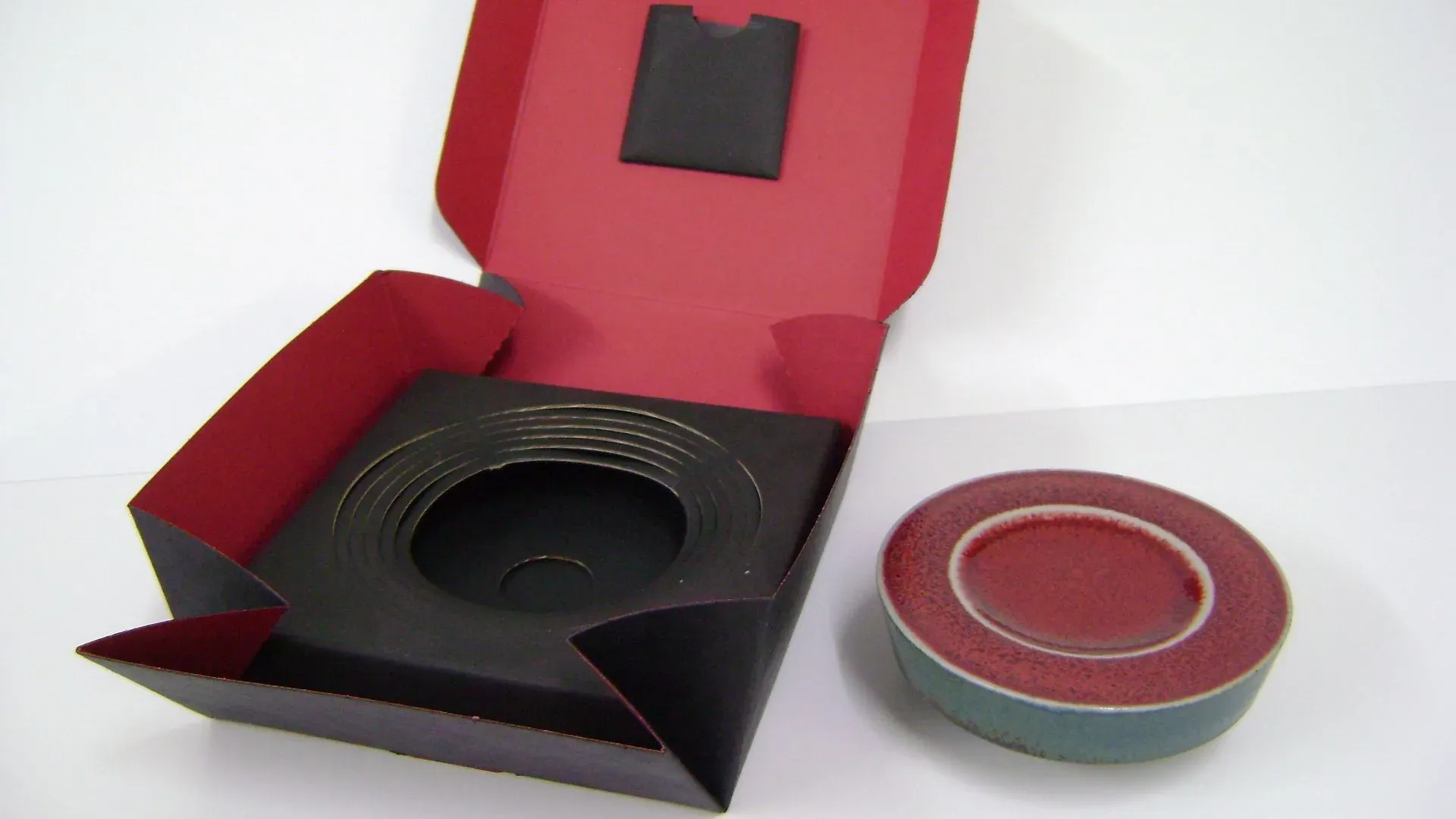 Offene rot-schwarze Schachtel mit einem Stapel Scheiben, neben einem passenden rot-grünen runden Behälter.
