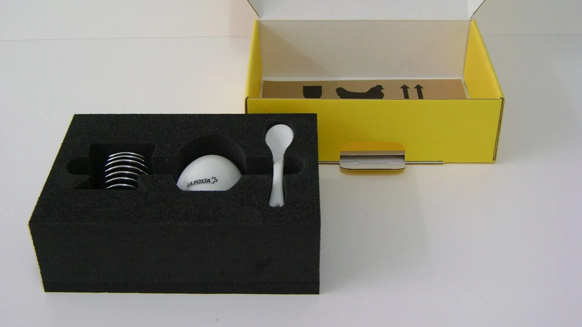 Eierbecher-Set aus schwarzem Schaumstoff, gelbe Box im Hintergrund. Enthält Ei, Löffel und Spiralhalter.