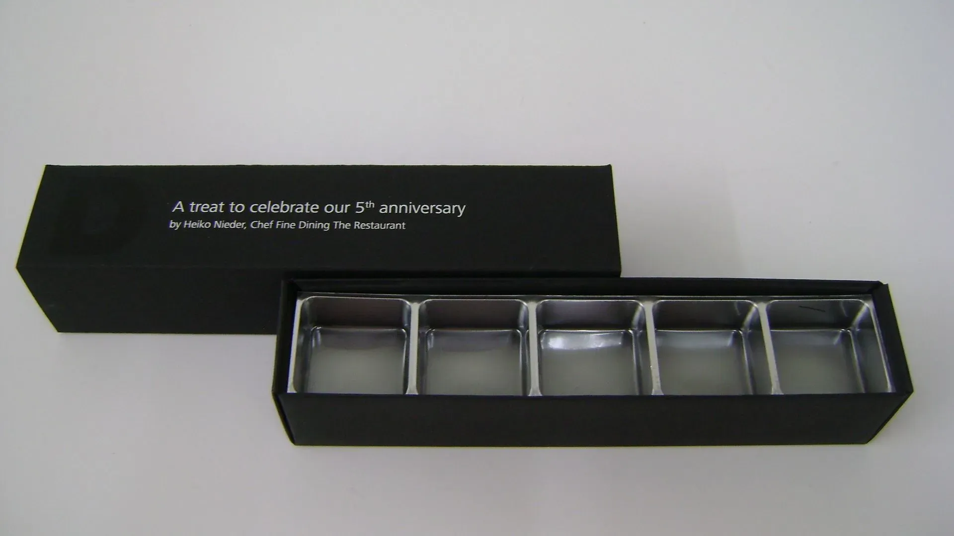 Schwarze Geschenkbox mit silberfarbenen Fächern, teilweise geöffnet, sodass fünf leere quadratische Felder sichtbar sind. Text auf dem Deckel.