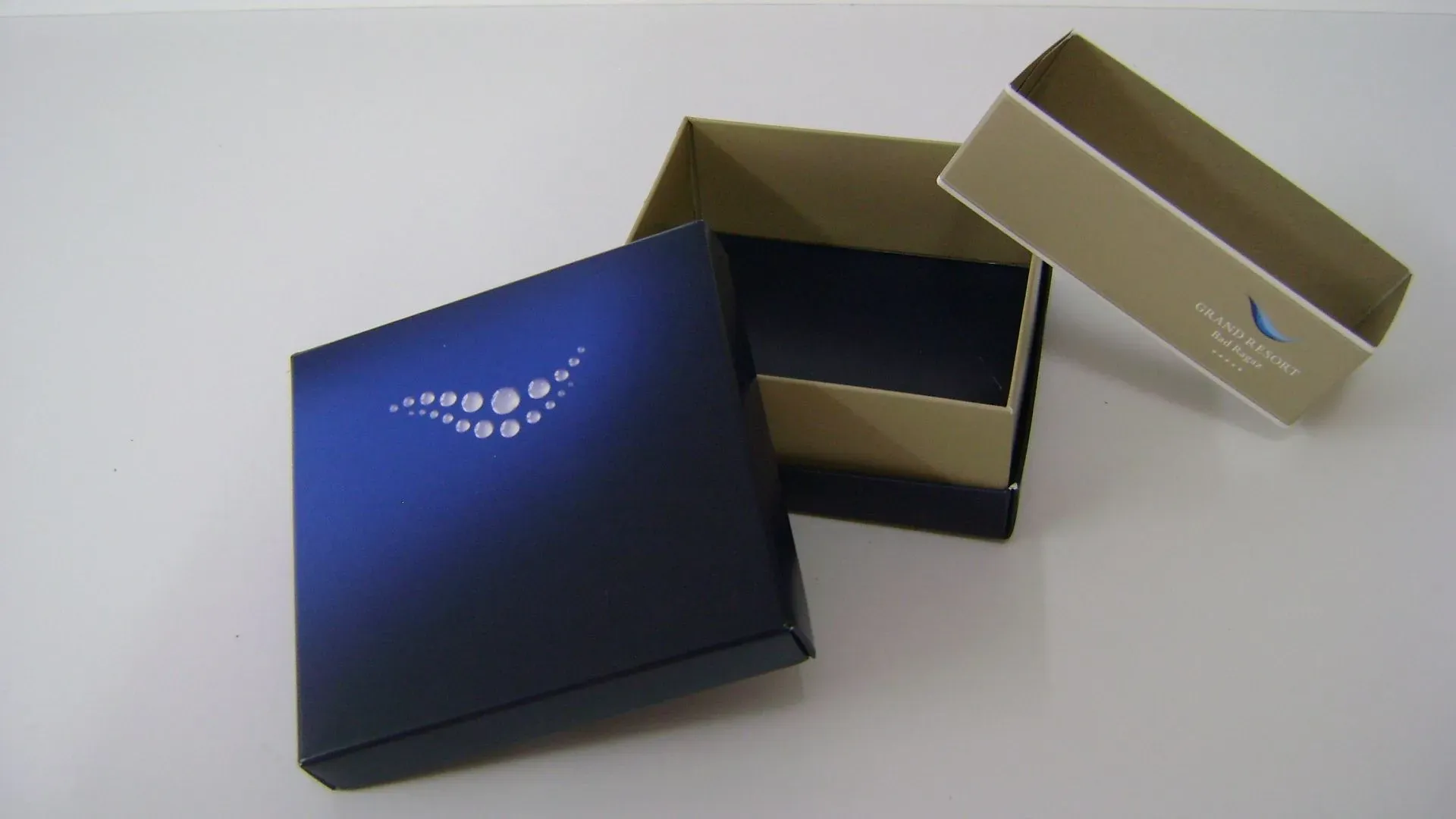 Eine quadratische Schachtel mit blauem Deckel und eine beige Schachtel, geöffnet auf einer weißen Fläche.