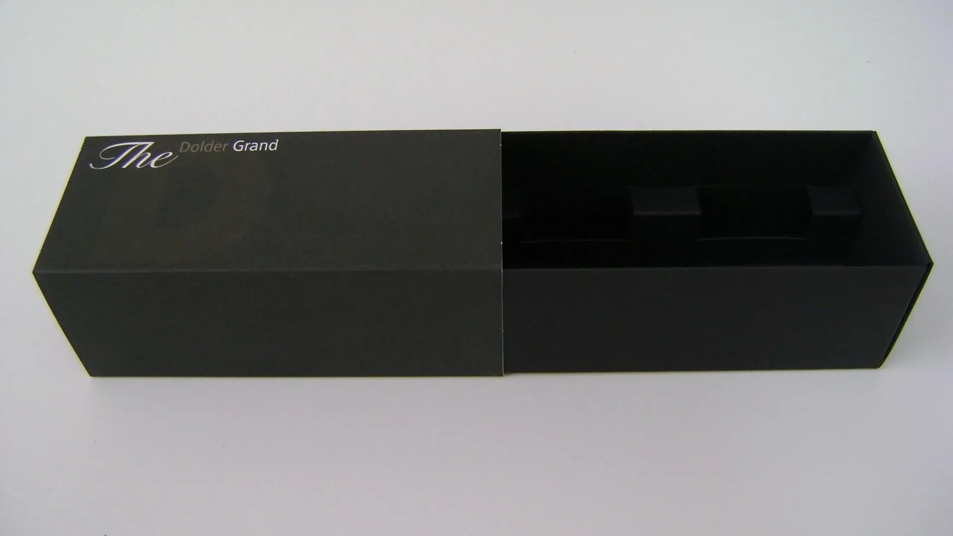 Schwarze rechteckige Schachtel, teilweise geöffnet, mit dunklem Innenraum mit Produktformen. Auf der Oberseite ist „The Awe“ aufgedruckt.