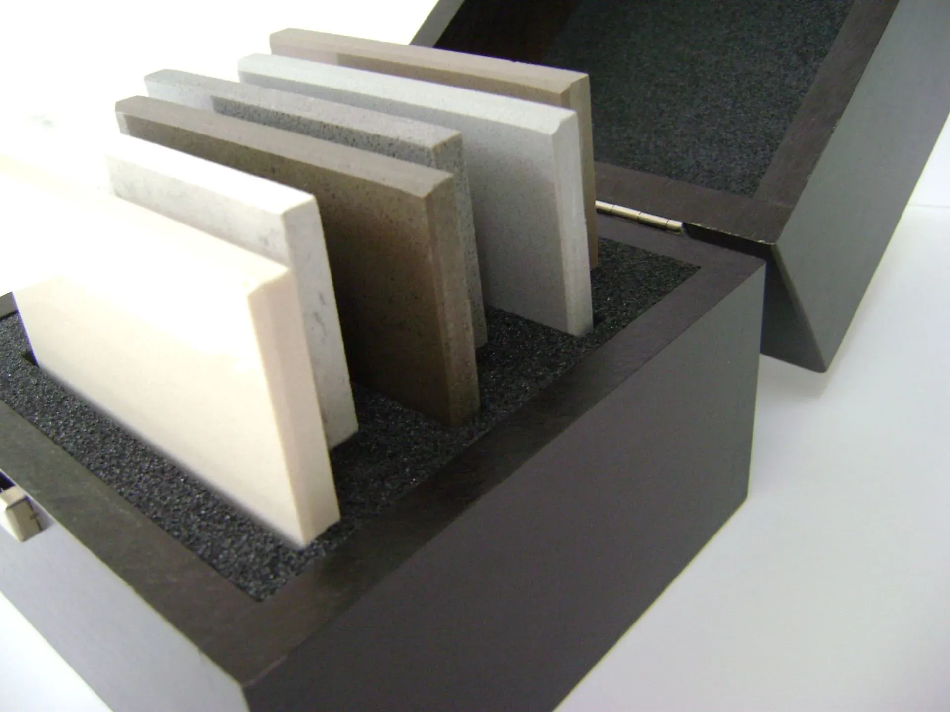 Fliesenmuster in einer dunklen Holzbox mit schwarzen Schaumstoffeinlagen. Verschiedene Farben und Texturen.