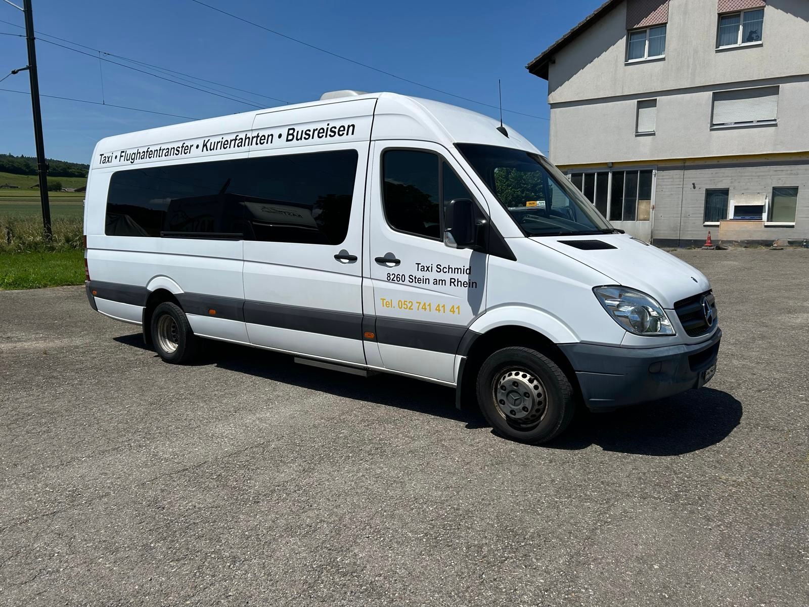 Ein blauer Mercedes-Benz-Transporter mit der Aufschrift „Taxi Schwyz“ parkt auf einer Kopfsteinpflasterstrasse. Eine Person sitzt am Steuer.