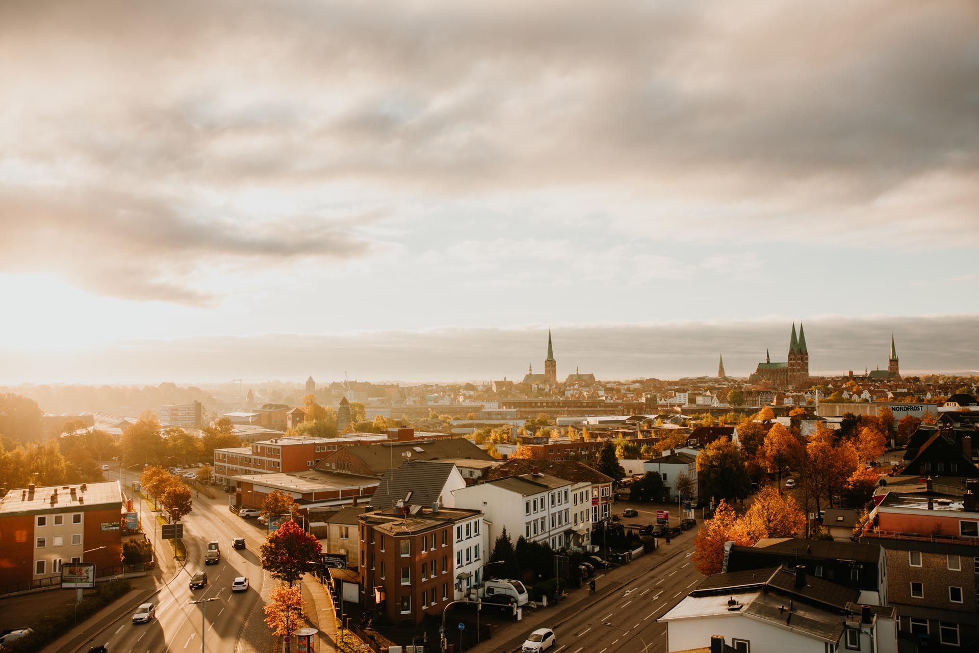 Sonnenbeschienene Stadtkulisse mit herbstlich gefärbten Bäumen, Dächern und einer fernen Kathedrale unter bewölktem Himmel