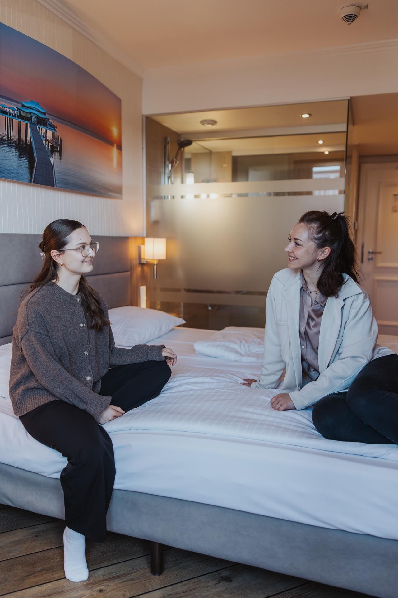 Zwei Frauen sitzen einander gegenüber auf einem Bett in einem warmen Hotelzimmer und unterhalten sich.