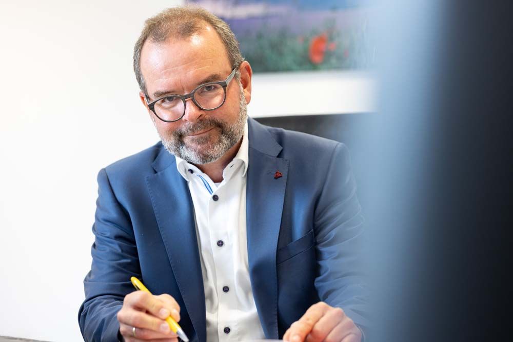 Mann mit Brille in blauem Anzug, der einen Stift hält, lächelt drinnen in die Kamera.