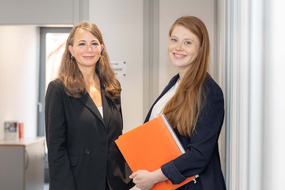 Zwei Frauen in Geschäftskleidung, eine davon mit einem orangefarbenen Ordner, stehen lächelnd in einem Flur.