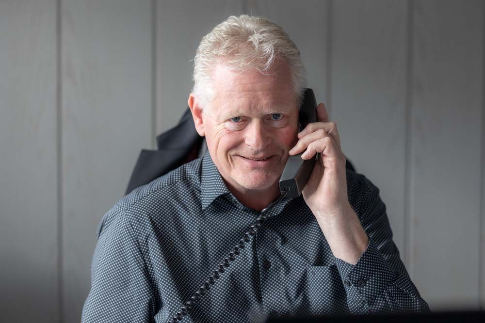 Älterer Mann mit grauem Haar, lächelnd, an einem Festnetztelefon in einem Büro, trägt ein gemustertes Hemd.