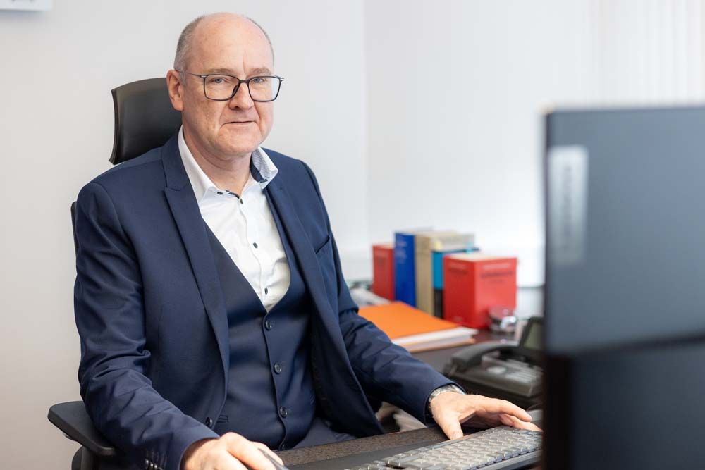 Mann im Anzug sitzt am Schreibtisch, tippt und lächelt. Büroumgebung, Bücher im Hintergrund.