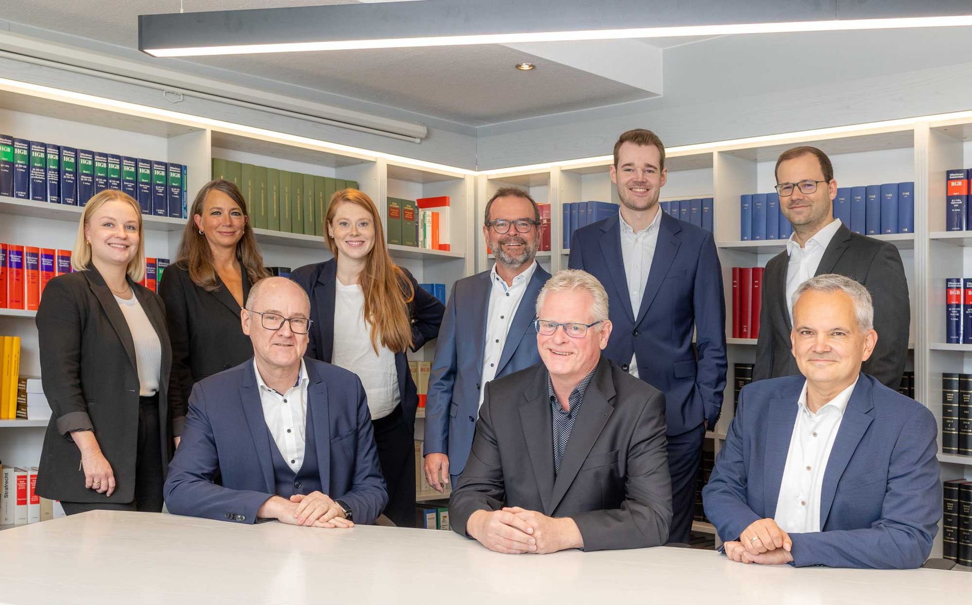 Eine Gruppe von Anwälten unterschiedlichen Alters posiert in einem Büro mit Bücherregalen. Einige sitzen, andere stehen.