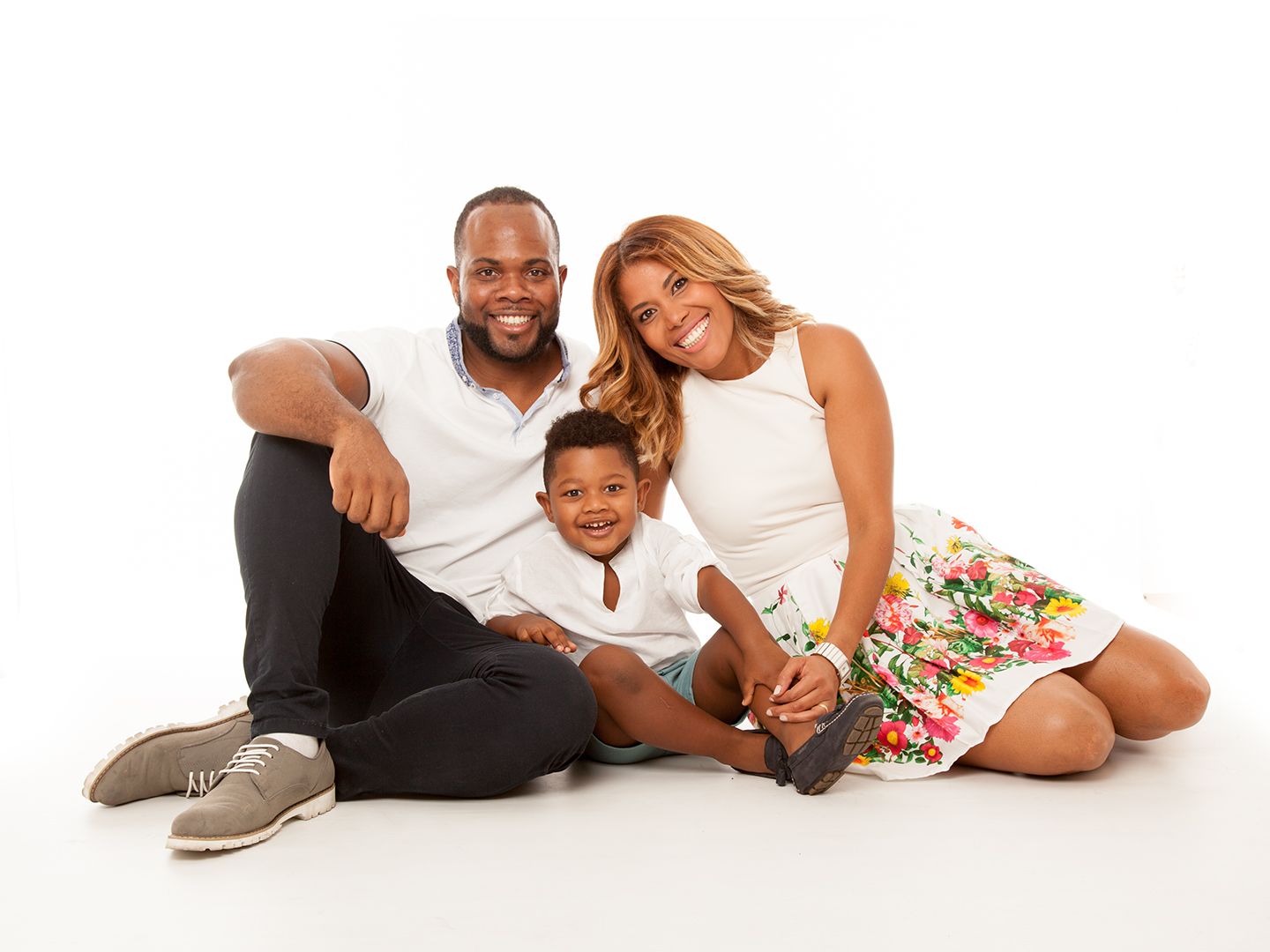 Familia de tres sonrientes, sentada sobre un fondo blanco; hombre con polo, mujer con vestido de flores, niño con camisa blanca.