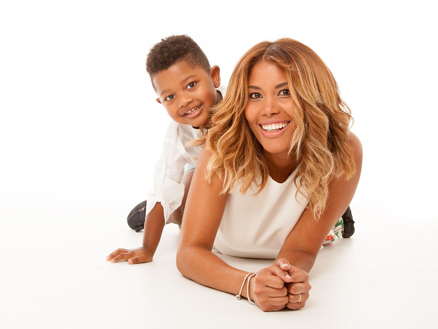 Mujer y niño sonriendo a la cámara. La mujer está tumbada, el niño está apoyado en su espalda. Fondo blanco.