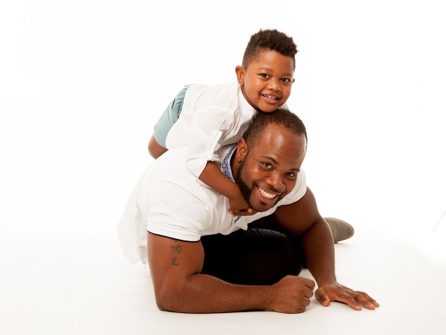 Hombre en el suelo con un niño a su espalda, ambos sonriendo, fondo blanco.