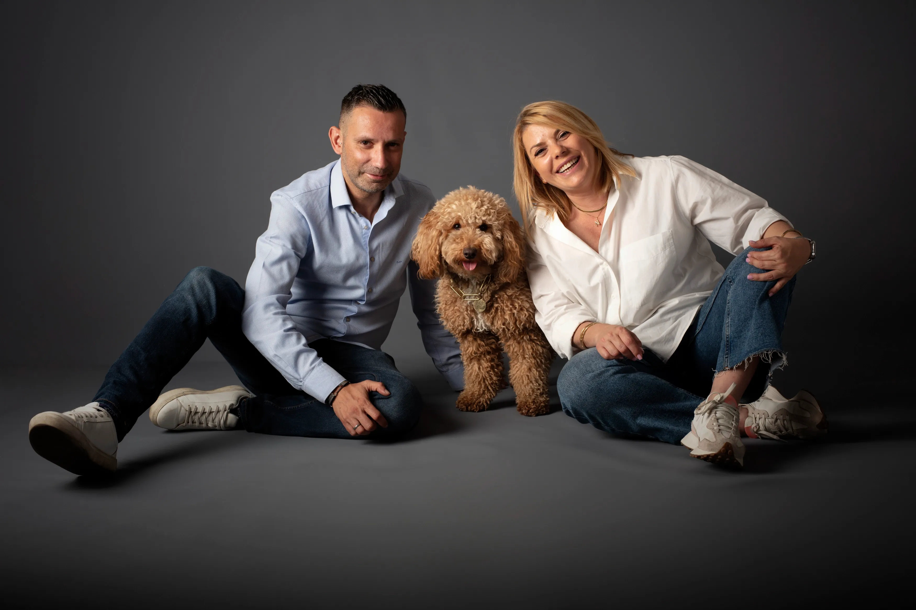 Un hombre y una mujer están sentados con un perro goldendoodle; todos sonríen contra un fondo gris.