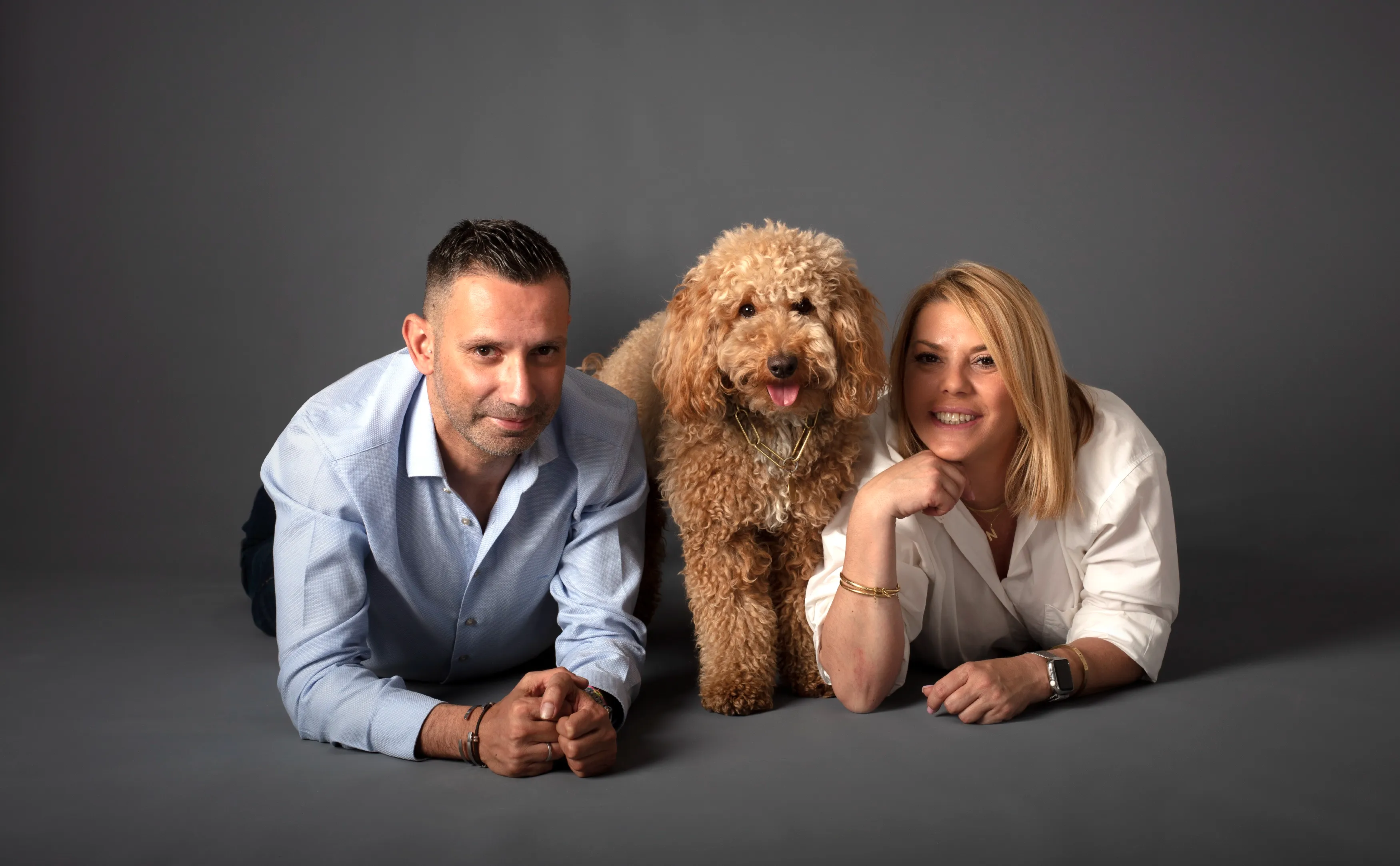 Una pareja y su perro posan sobre un fondo gris. El perro está de pie entre ellos, sonriendo.
