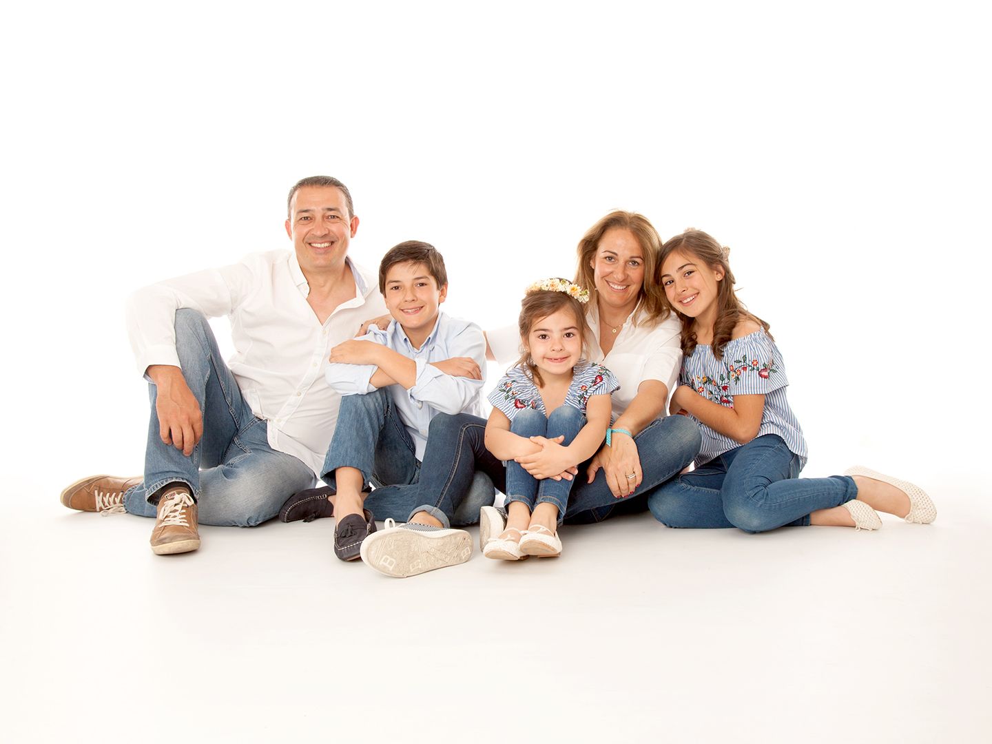 Una familia de cinco personas sonríe, sentada muy junta sobre una superficie blanca. Todos visten vaqueros y camisetas blancas.