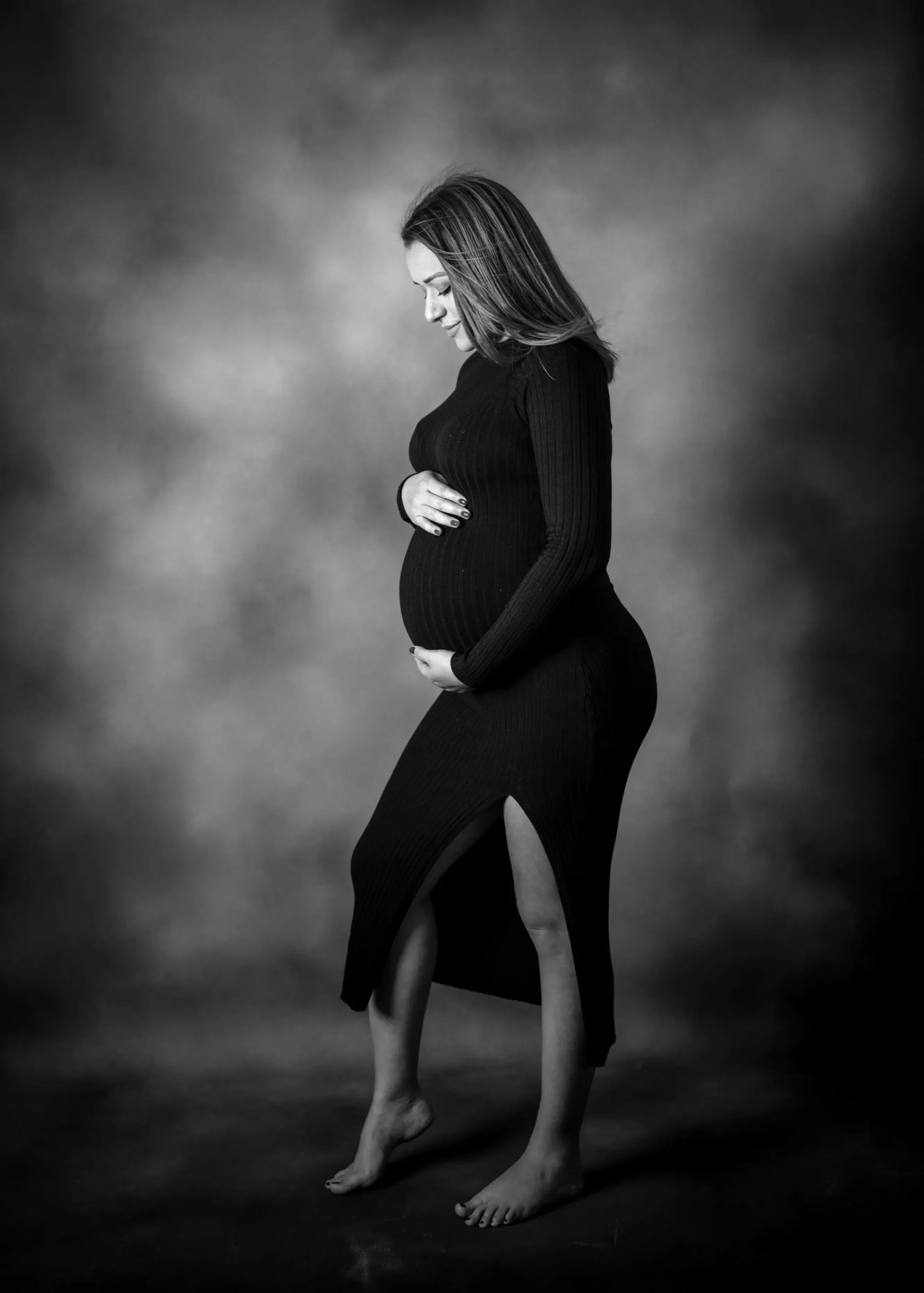 Mujer embarazada con vestido negro, mano sobre el vientre, perfil lateral, mirando hacia abajo. Fondo de estudio.