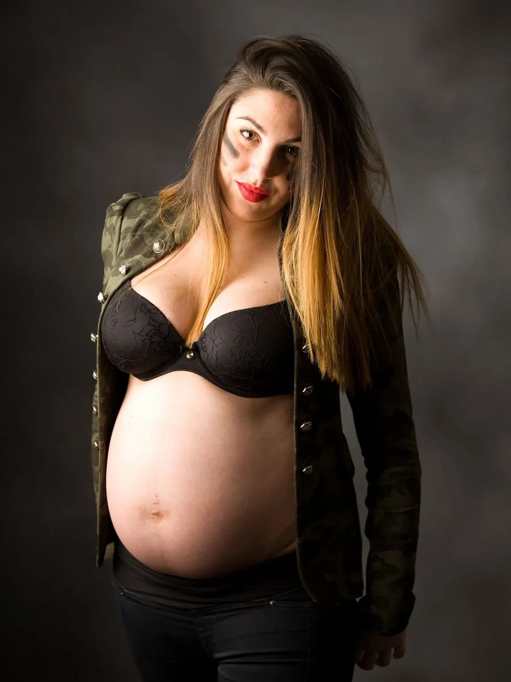 Mujer embarazada con sujetador negro, chaqueta de camuflaje, sonriendo con pintalabios rojo, mano sobre el vientre.