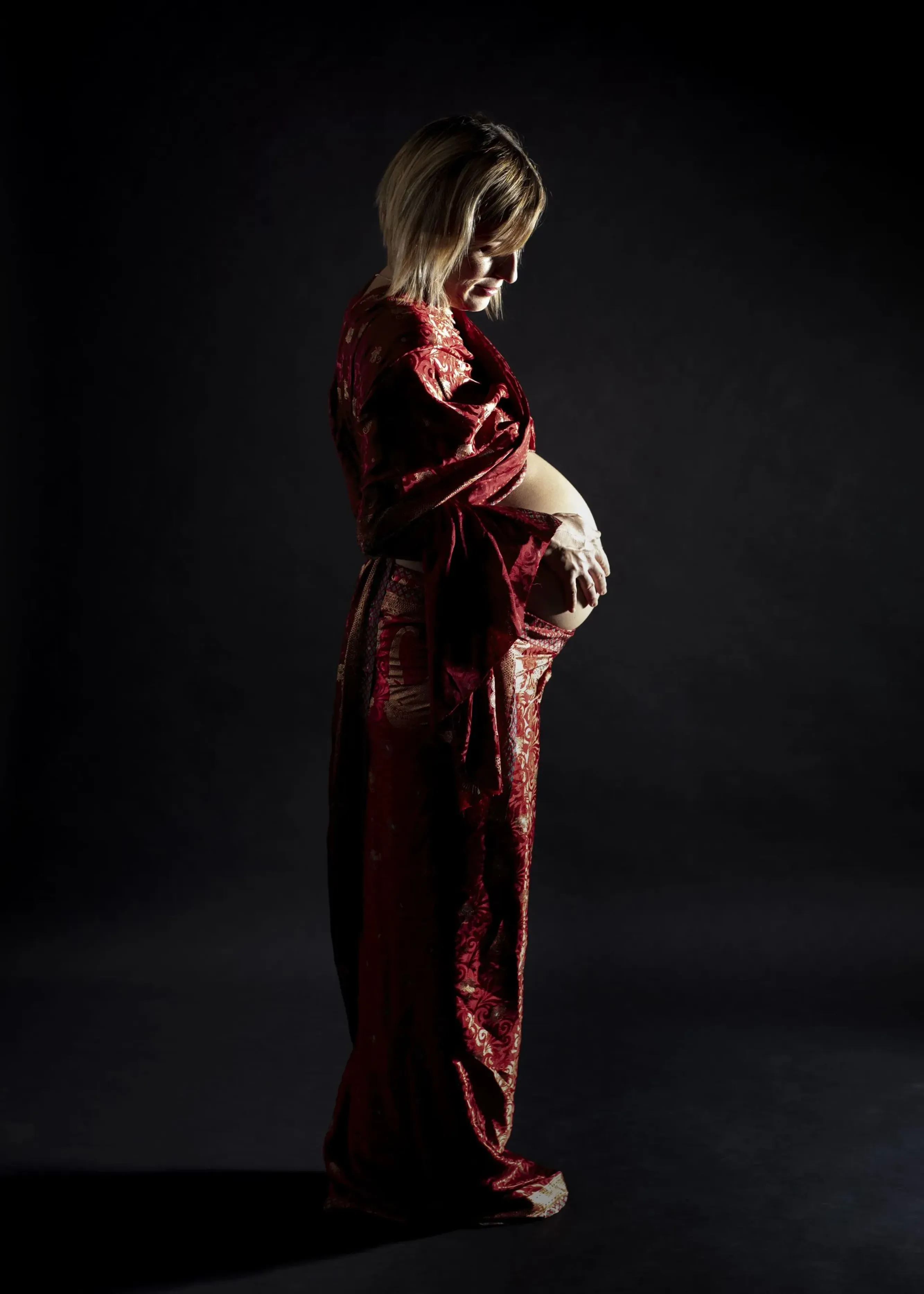 Mujer embarazada con un vestido rojo de lentejuelas, sosteniendo su abdomen; fondo oscuro.