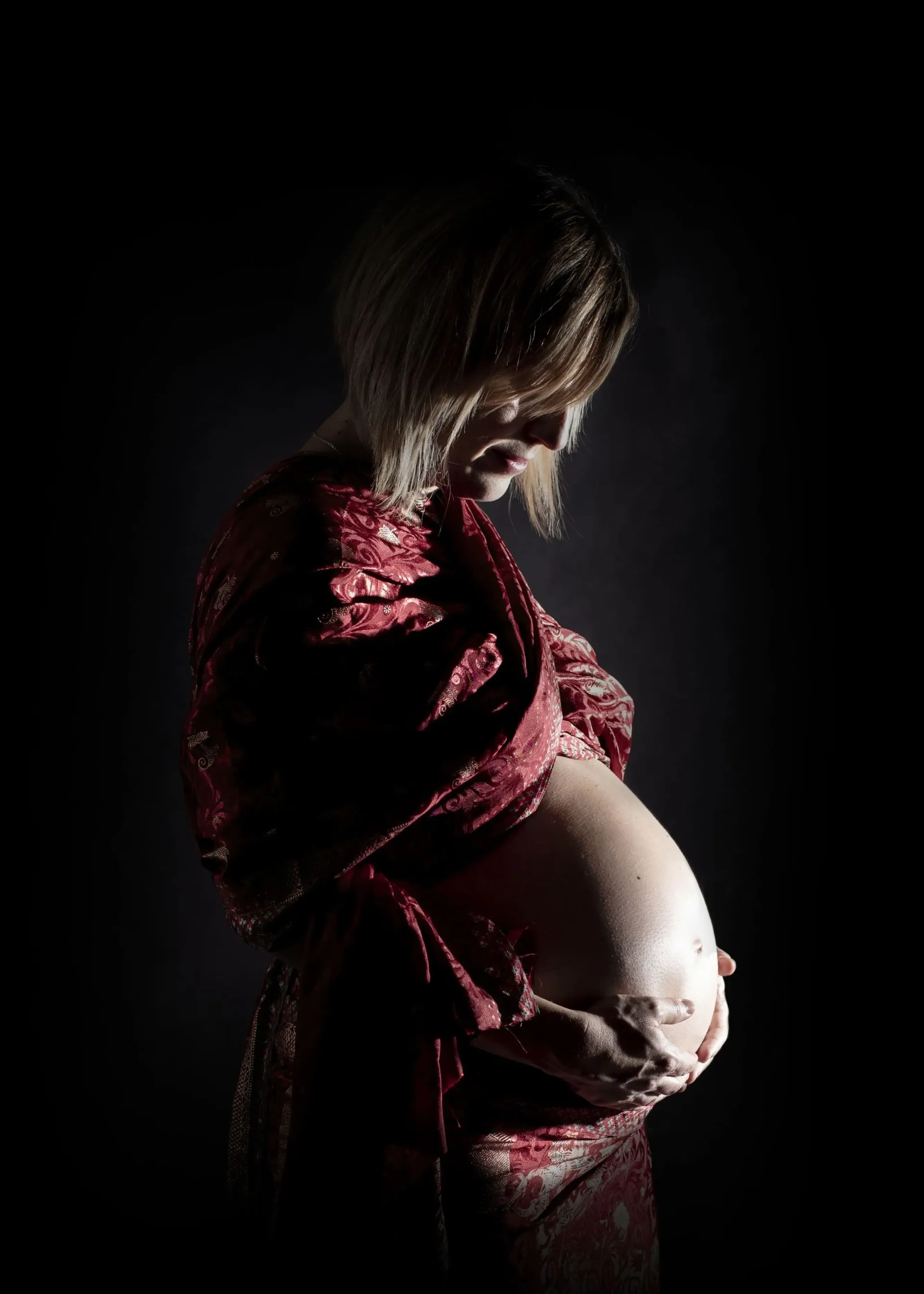 Mujer embarazada acariciando su vientre, iluminada contra un fondo oscuro, vestida con una bata roja estampada.