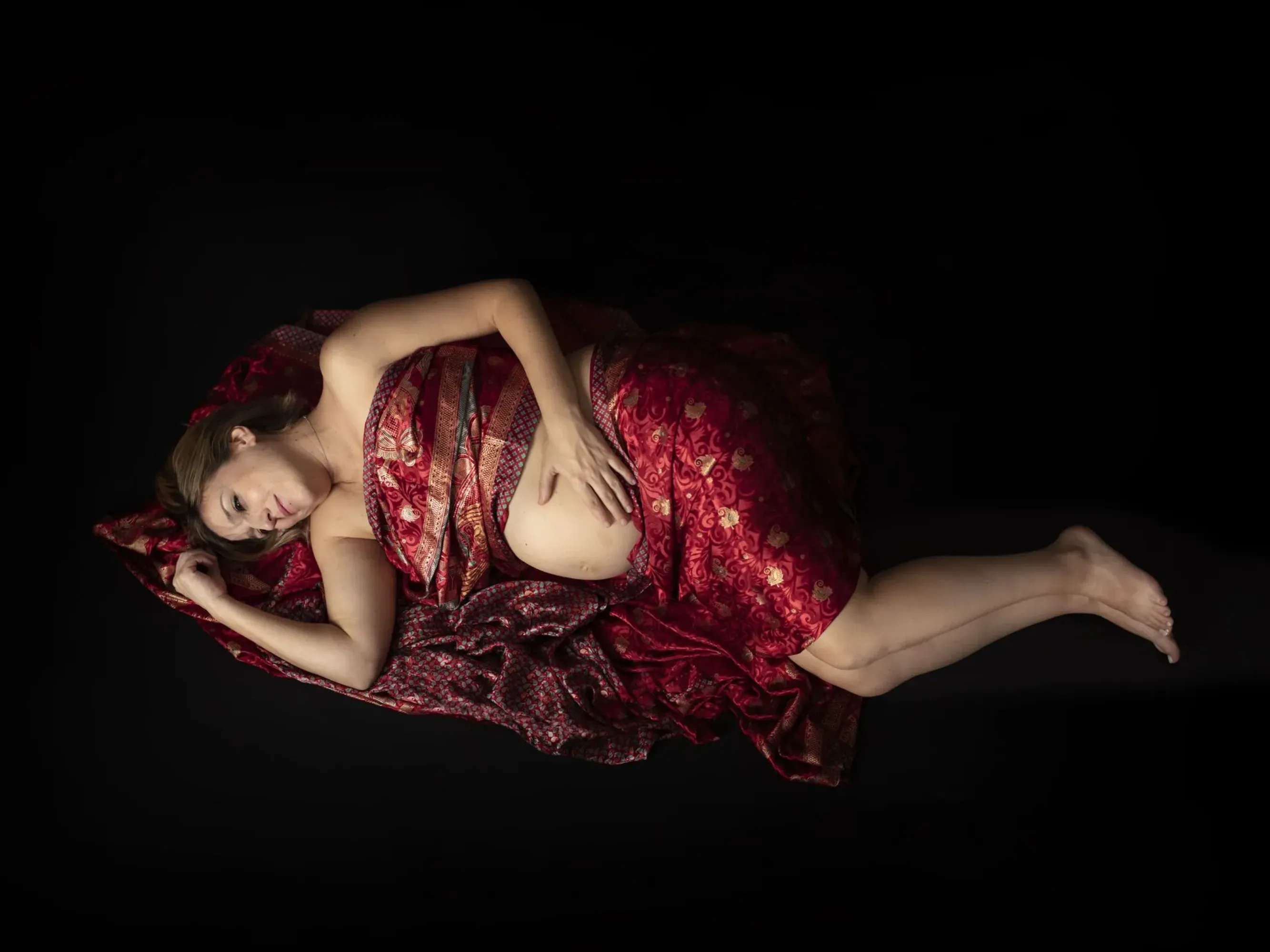 Mujer vestida de rojo, recostada, con la mano sobre el abdomen, fondo oscuro.