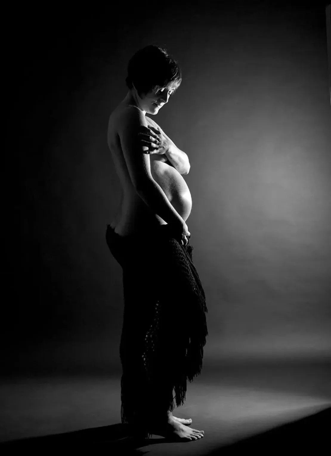 Mujer embarazada, de perfil, con el vientre prominente, la mano sobre el pecho, parcialmente cubierta. Estudio oscuro, con sombras.