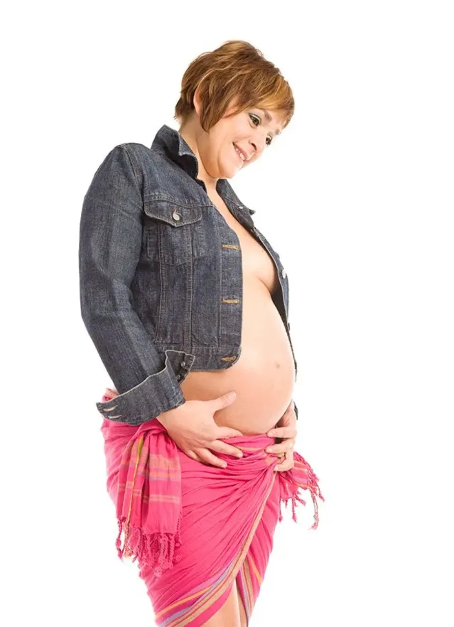 Mujer embarazada con chaqueta vaquera y pareo rosa, mirando hacia abajo a su vientre.