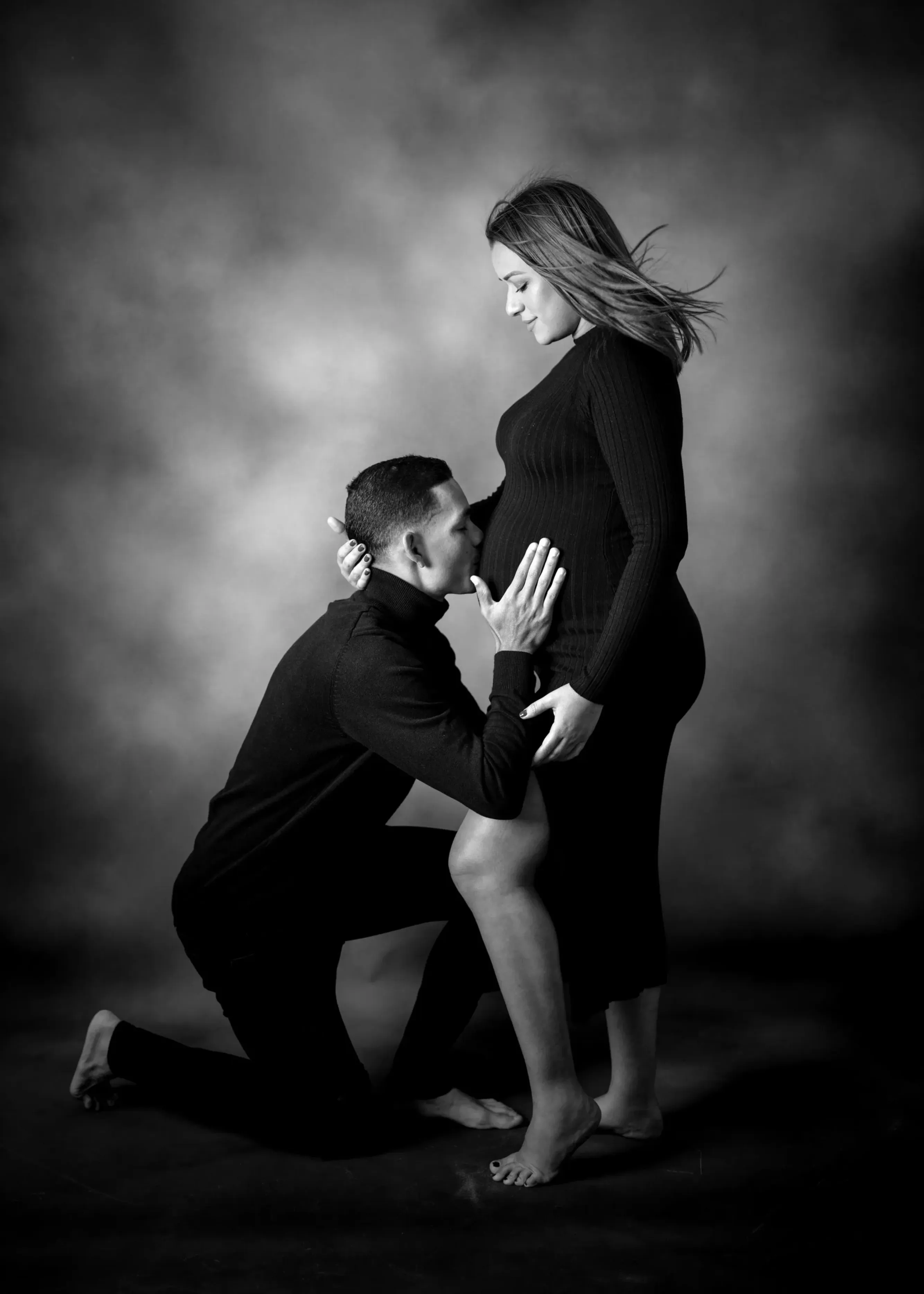 Hombre arrodillado besando el vientre de una mujer embarazada; ambos vestidos de negro, en un estudio.