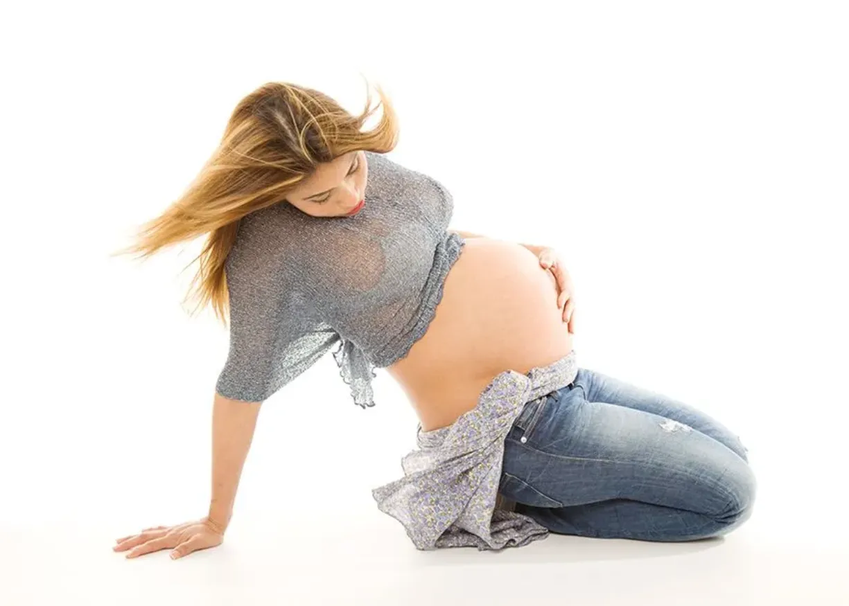 Mujer embarazada arrodillada, vestida con una camisa azul y pantalones vaqueros, tocándose el vientre.