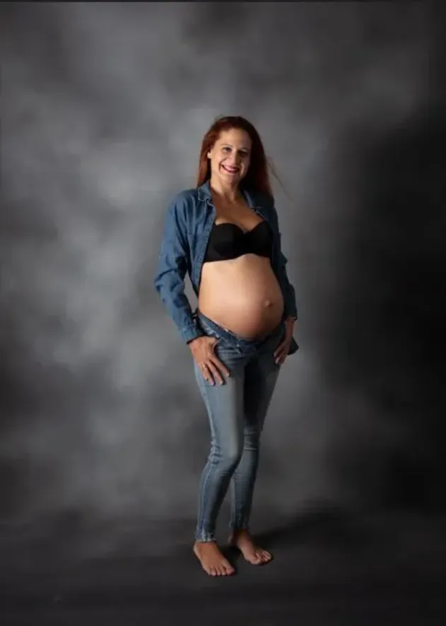 Mujer embarazada en vaqueros y camisa vaquera, abdomen descubierto, sonriendo, fondo de estudio.