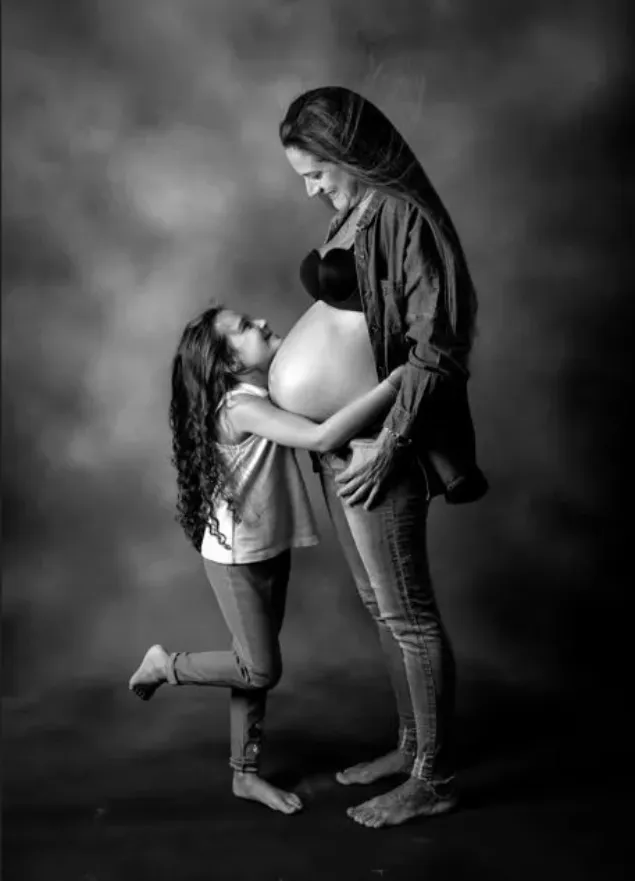 Una mujer embarazada abraza a una niña pequeña; ambas sonríen y ella sostiene el vientre de la mujer. Fotografía de estudio en blanco y negro.