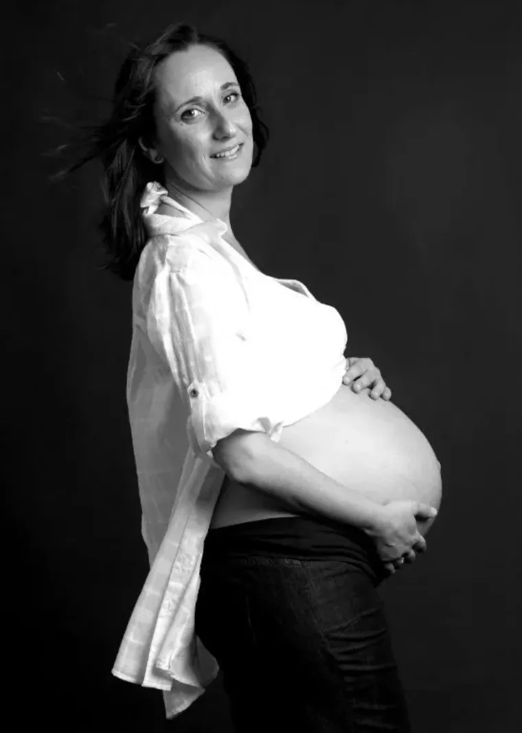Mujer embarazada con camisa blanca y pantalones oscuros, sosteniendo su vientre, sonriendo, fondo oscuro.