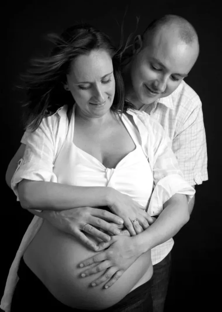Mujer embarazada y hombre abrazados, con las manos sobre el vientre, sonriendo; retrato de estudio en blanco y negro.