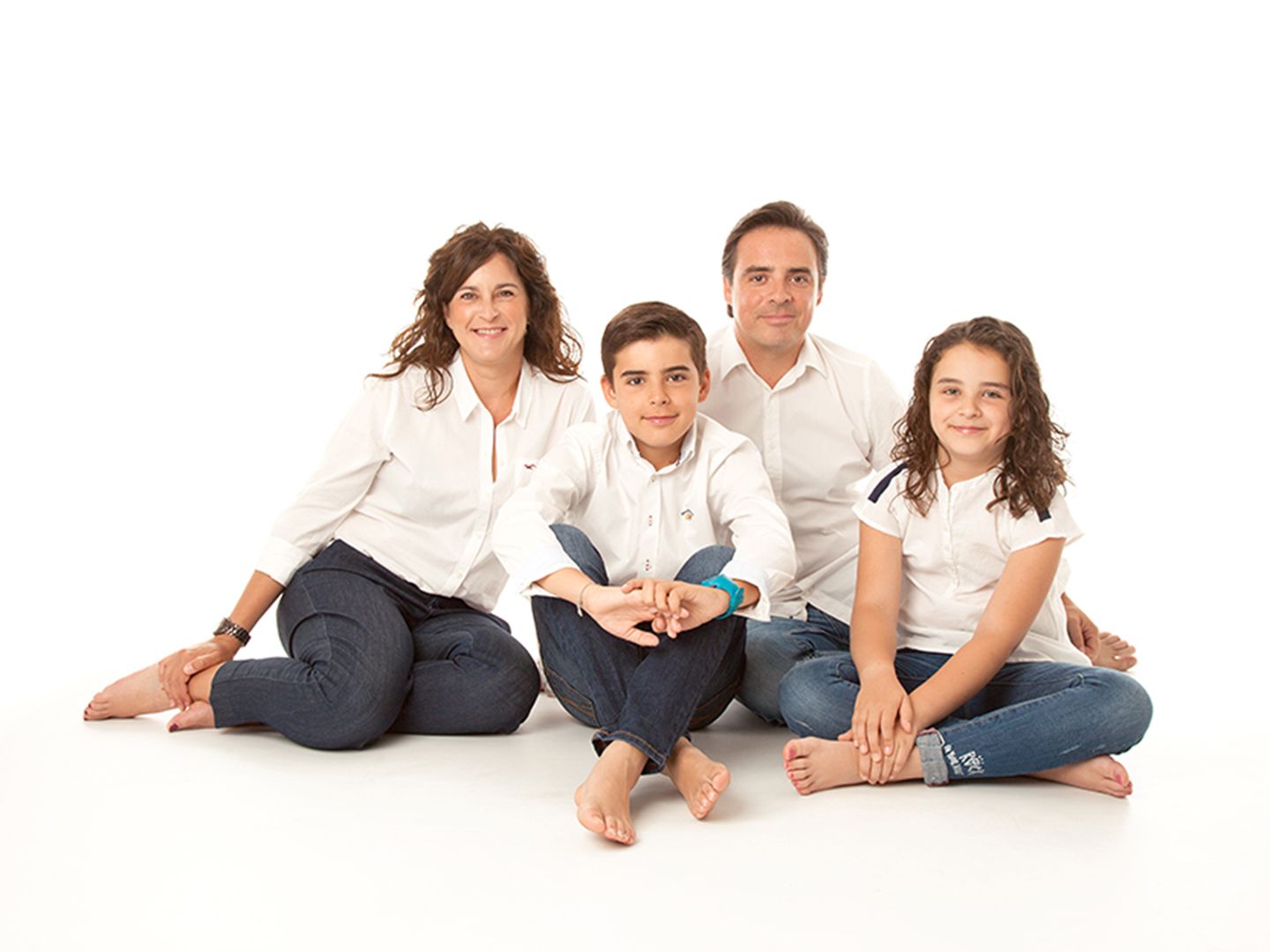 Familia de cuatro integrantes, sonrientes, vestidos con camisas blancas y pantalones vaqueros azules, sentados sobre fondo blanco.