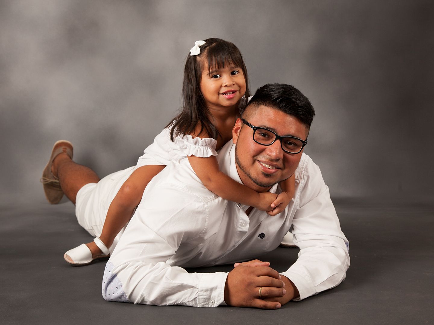 Hombre vestido de blanco tumbado en el suelo con una niña pequeña a su espalda. Ambos sonríen.