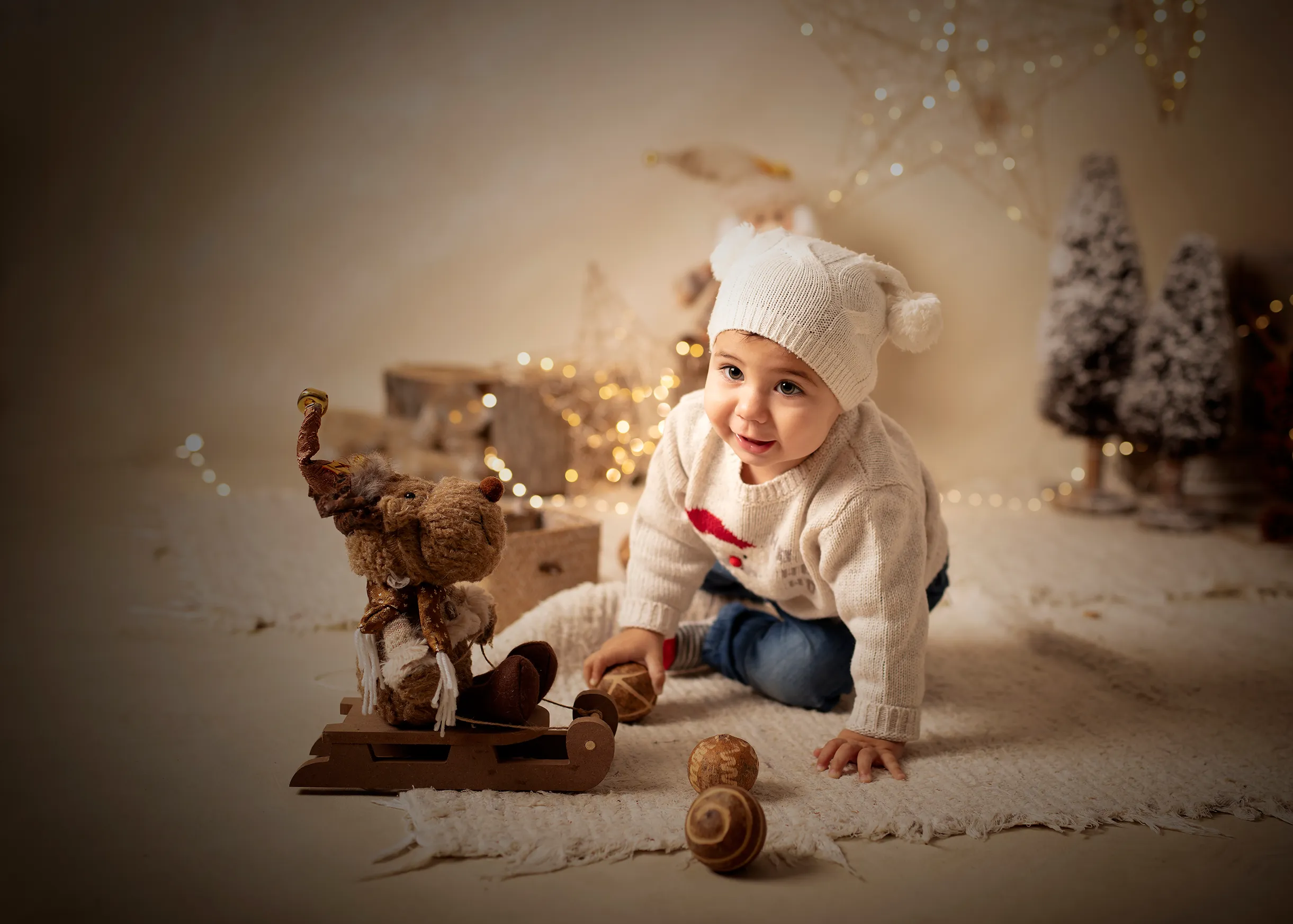 Un bebé con gorro y suéter de lana juega con juguetes de madera en un ambiente invernal con luces y arbolitos.